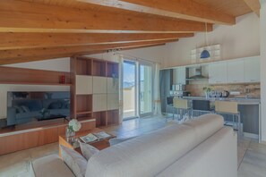 A modern living room with a large flat-screen TV, a sofa, and a coffee table. There is a sliding glass door leading to a balcony with a view of the mountains. The kitchen is visible in the background with white cabinets and a blue pendant light. - Magic Lake View in Lierna (Lierna)