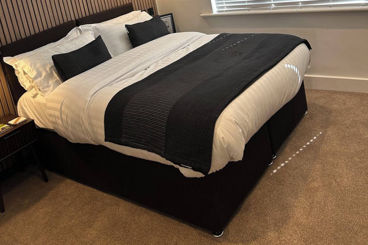A modern bedroom with a large bed, a wooden slat headboard, a bedside table, and a window with blinds.