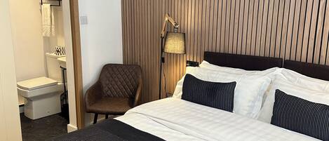A hotel room with a large bed, a bedside lamp, a chair, a bathroom visible through an open door, and a wooden panel wall.