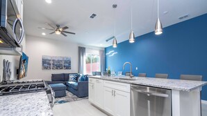 Una cocina moderna con gabinetes blancos, lavavajillas de acero inoxidable y encimera de granito. Presenta una pared de acento azul, ventilador de techo e iluminación colgante sobre la isla de la cocina.