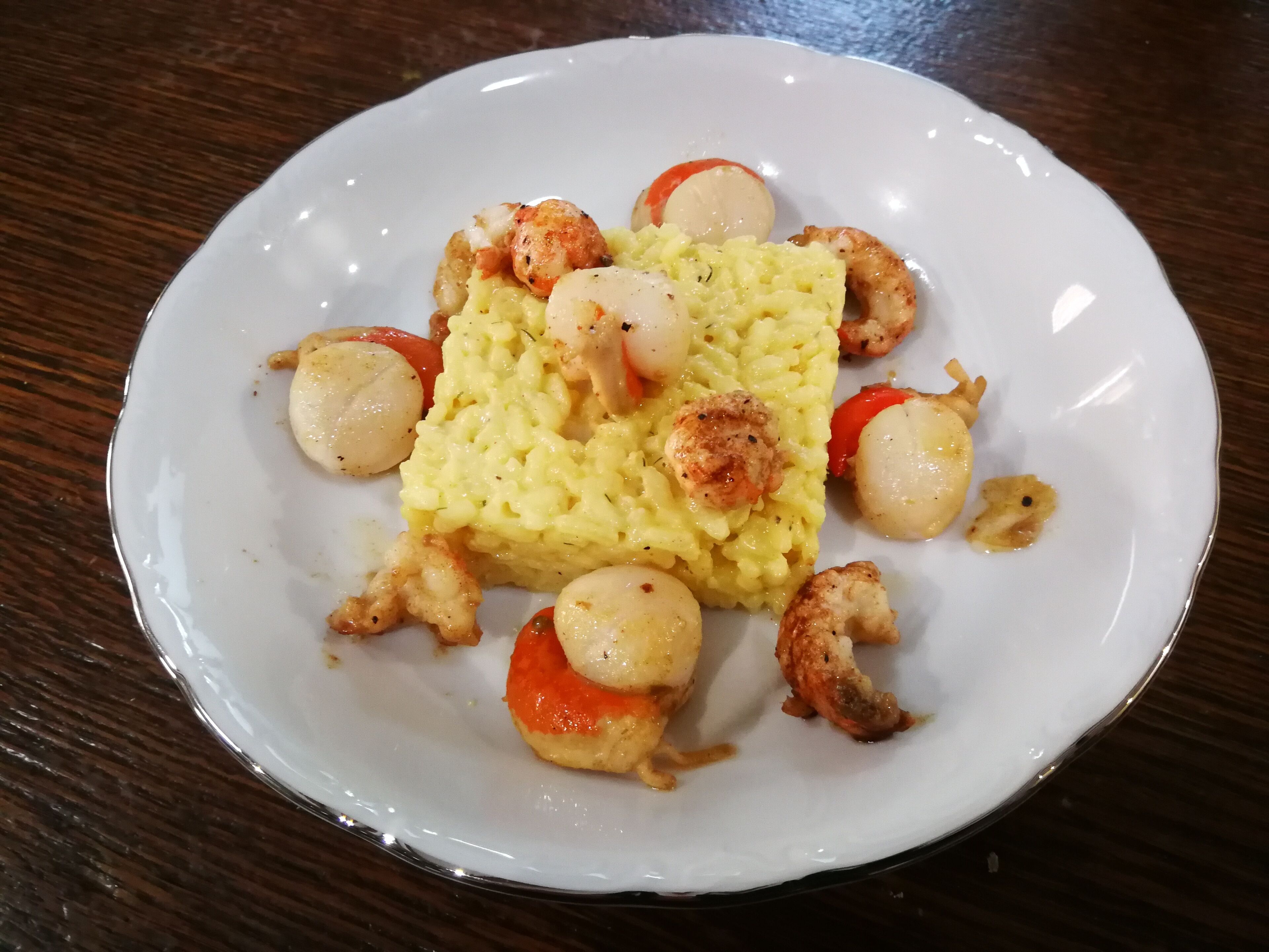 Um prato branco com risoto amarelo, vieiras e tomates cereja sobre uma mesa de madeira escura.