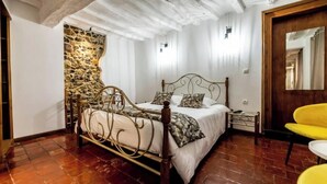 Une chambre avec un mur en pierre, un lit avec un cadre en métal, une porte en bois et une chaise jaune.