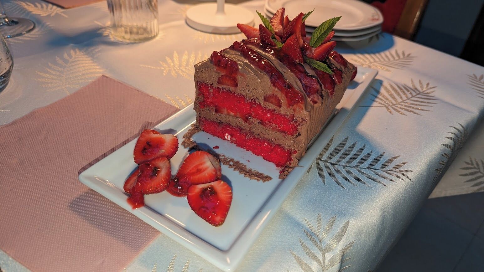 A slice of layered cake with strawberries on a plate, placed on a table with a patterned tablecloth.