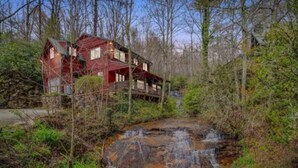 Un chalet rustique doté d’une terrasse en bois, entouré d’une forêt dense et traversé par un petit ruisseau.