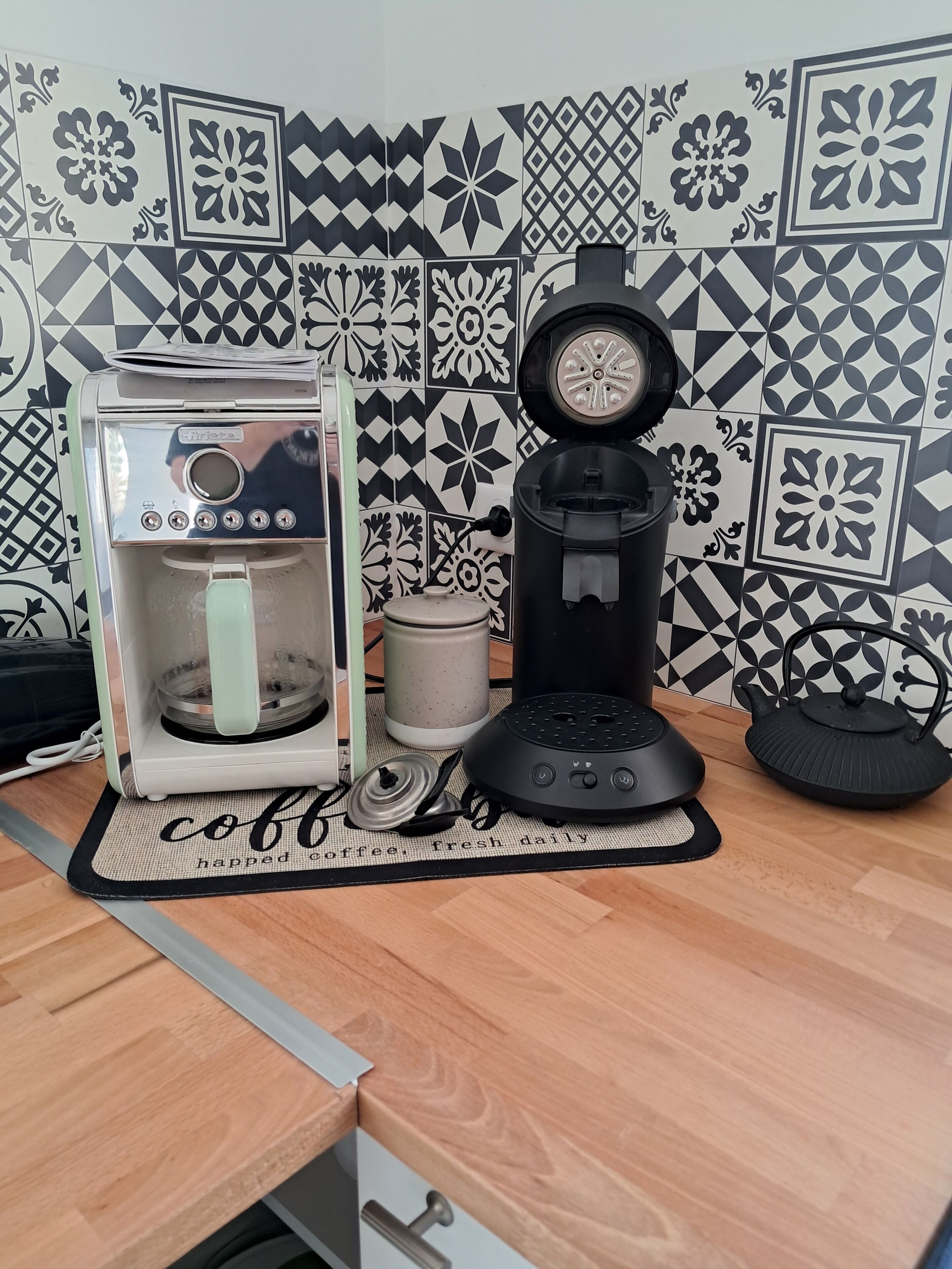 A coffee machine and a kettle on a kitchen counter with a coffee-themed mat.