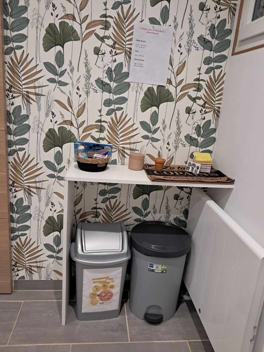 A kitchen area with a shelf holding a basket, a candle, and a tray of items. Below the shelf, there are two trash bins.