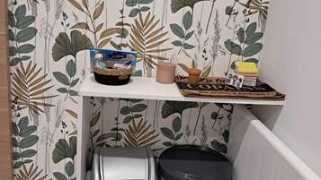 Una zona de cocina con un estante que sostiene una cesta, una vela y una bandeja con objetos. Debajo del estante, hay dos cubos de basura.