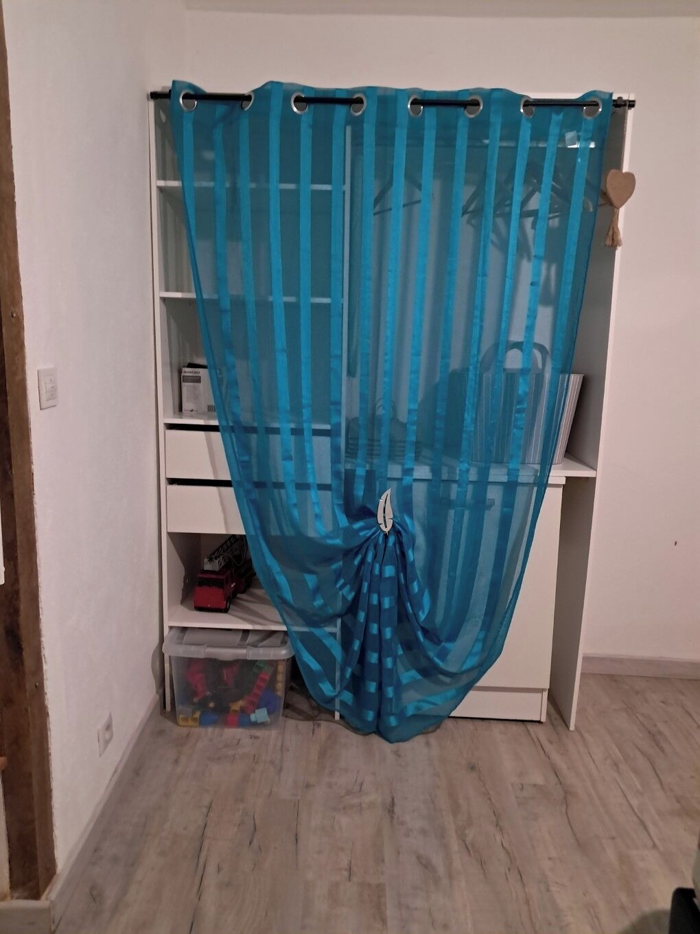 A room with a blue sheer curtain, a white shelving unit, and a plastic storage bin.