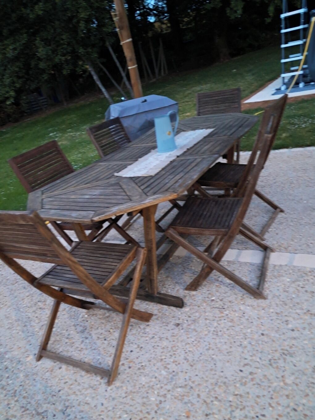 Une table en bois installée à l’extérieur, avec des chaises, un pichet bleu et une échelle en arrière-plan.