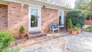 A brick house with a front door, a small porch with a chair and table, and potted plants. - Idlewild Studio: Scandinavian studio just 20min to uptown (Matthews)