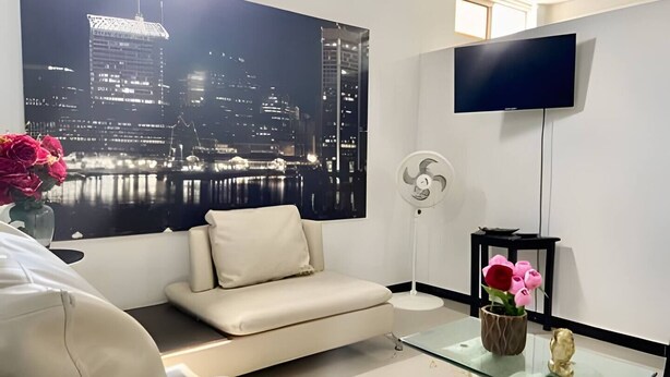 A modern living room with a sofa, a glass coffee table, a TV mounted on the wall, and a large window with a cityscape view.