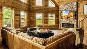 L’intérieur d’une cabane en rondins, avec une cheminée en pierre, un canapé en cuir et de grandes fenêtres donnant sur le paysage extérieur.