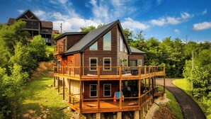 Une maison en bois à plusieurs étages, dotée d’un balcon, entourée de verdure et bordée d’une route sinueuse.