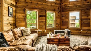 L’intérieur d’un chalet accueillant, avec un canapé en cuir, une table basse sur laquelle est posé un plateau de collations, et de grandes fenêtres offrant une vue sur la verdure et un bâtiment situé au loin.