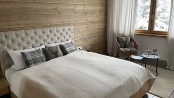 Ein Schlafzimmer mit einem hölzernen Kopfteil, einem Bett mit weißer Daunendecke, einem Sessel und einem kleinen Tisch mit zwei Weingläsern.