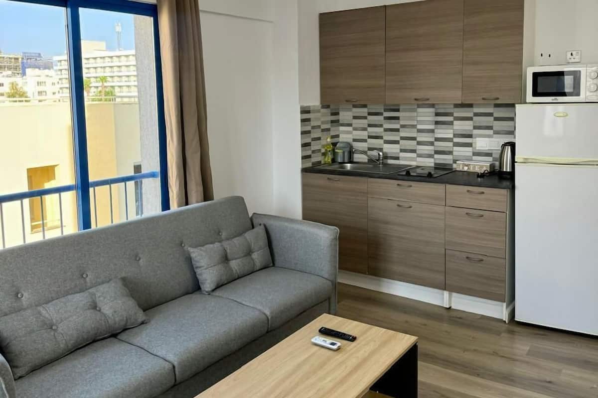 A modern living room with a grey sofa, a wooden coffee table, and a kitchen area with wooden cabinets and a microwave.