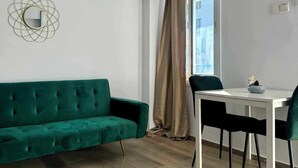 Una sala de estar moderna con un sofá verde, una mesa blanca con dos sillas y una ventana con cortinas color beige.
