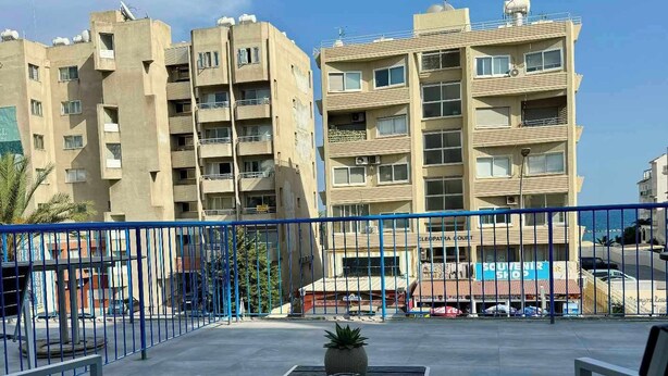 Un balcón con dos sillas y una mesita pequeña, con vistas a un edificio con varios balcones y una tienda de recuerdos.