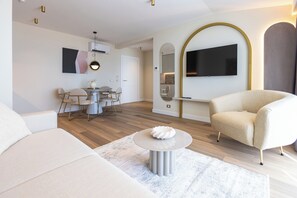 En moderne stue med sofa, rundt sofabord, spiseplads med stole, fjernsyn ophængt på væggen, og et køkkenområde synligt gennem en bue.