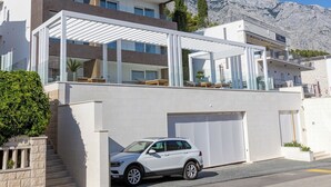 A modern two-story building with a white facade, a covered outdoor seating area, and a white car parked in front. - Luxus Villa M - Apartment 1 (Croatia)