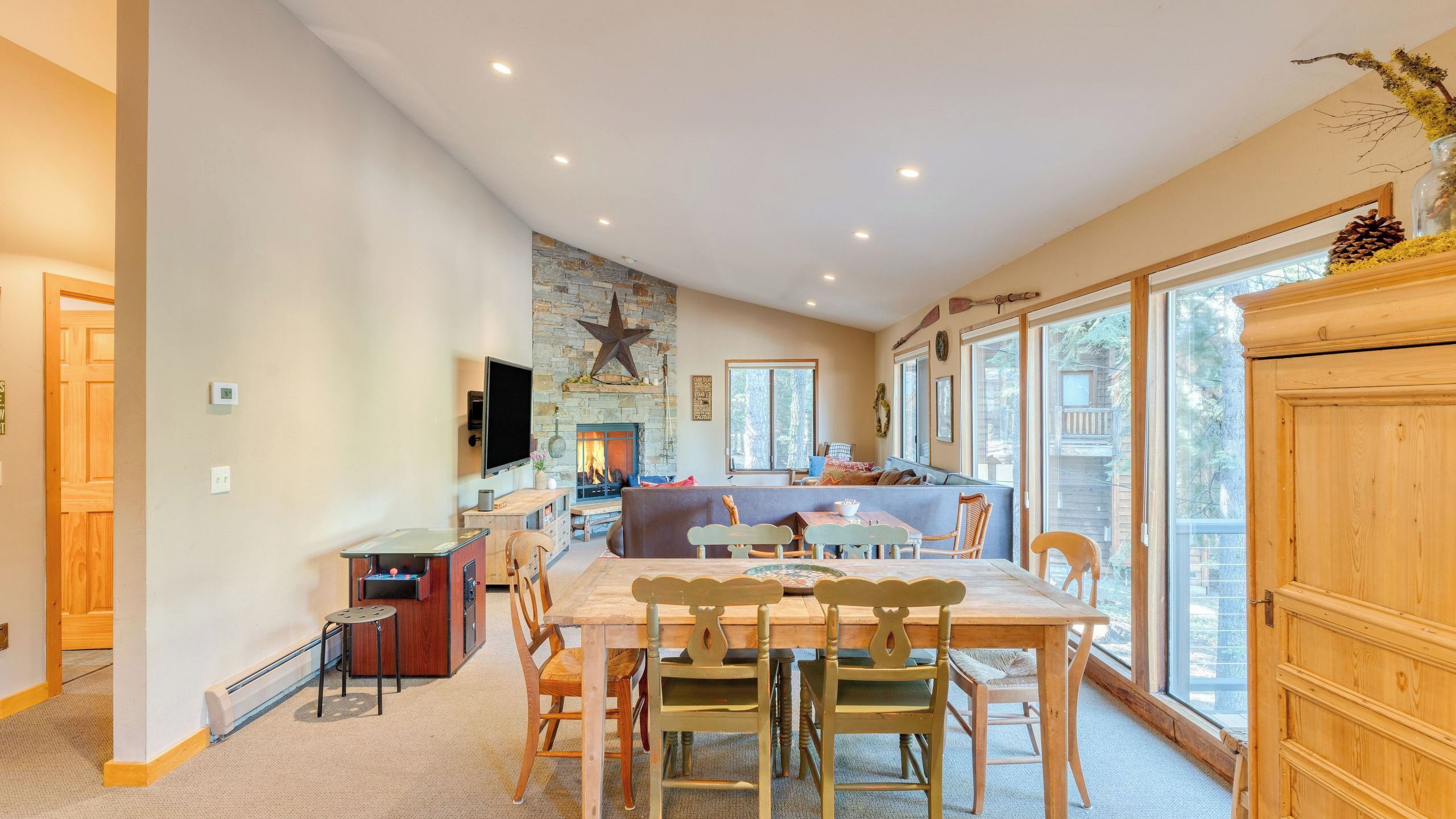 A spacious living area with a stone fireplace, a dining table with chairs, and large windows offering a view of the outdoors.