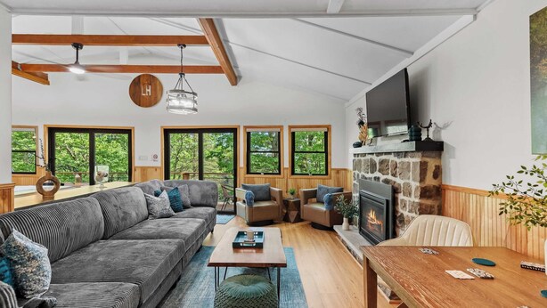 A spacious living room with a large sofa, a wooden coffee table, and a fireplace. There are multiple windows providing natural light and views of the outdoors.