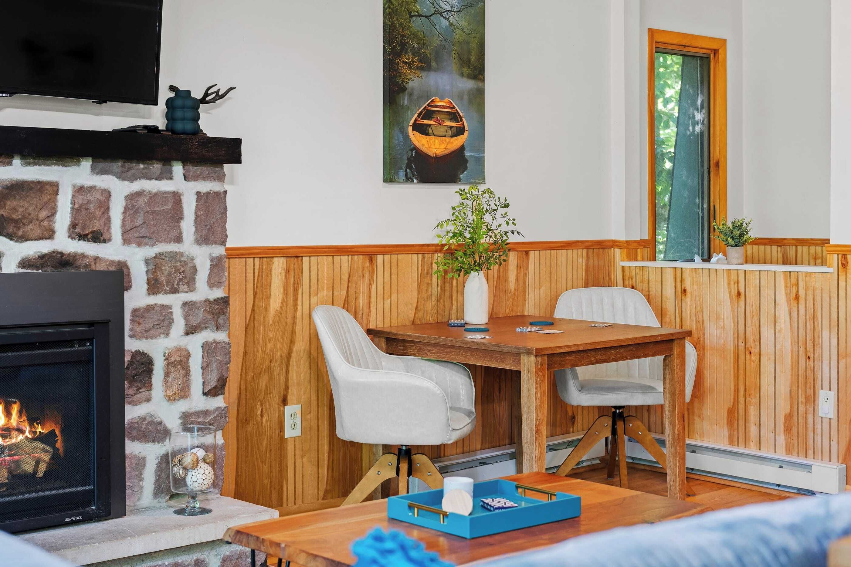 A cozy room with a stone fireplace, a wooden table with chairs, a TV mounted above the fireplace, and a window with a view of trees.