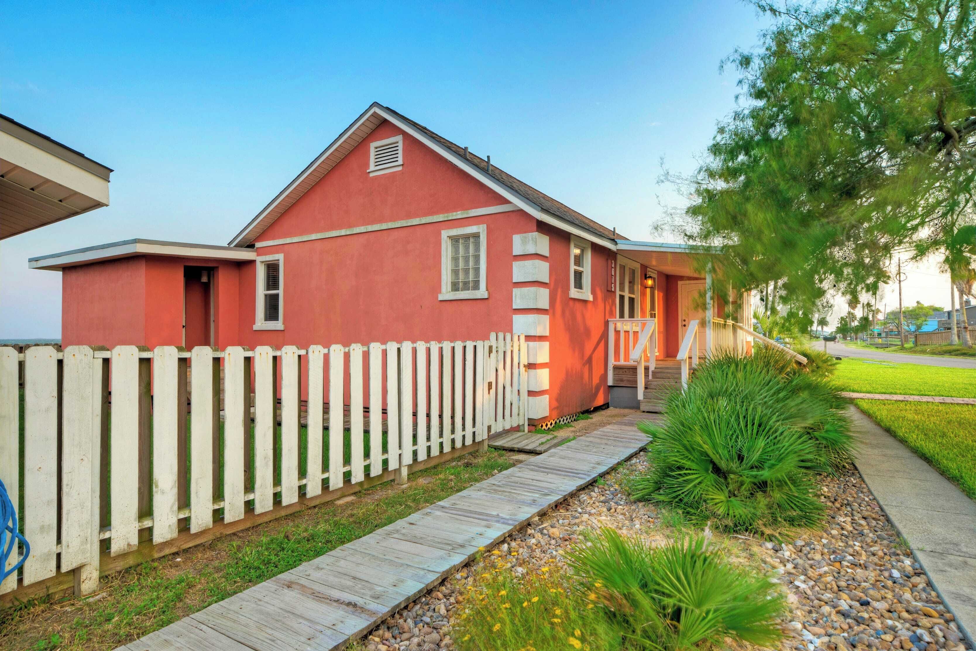 A red house with a white picket fence, a wooden walkway, and green landscaping.