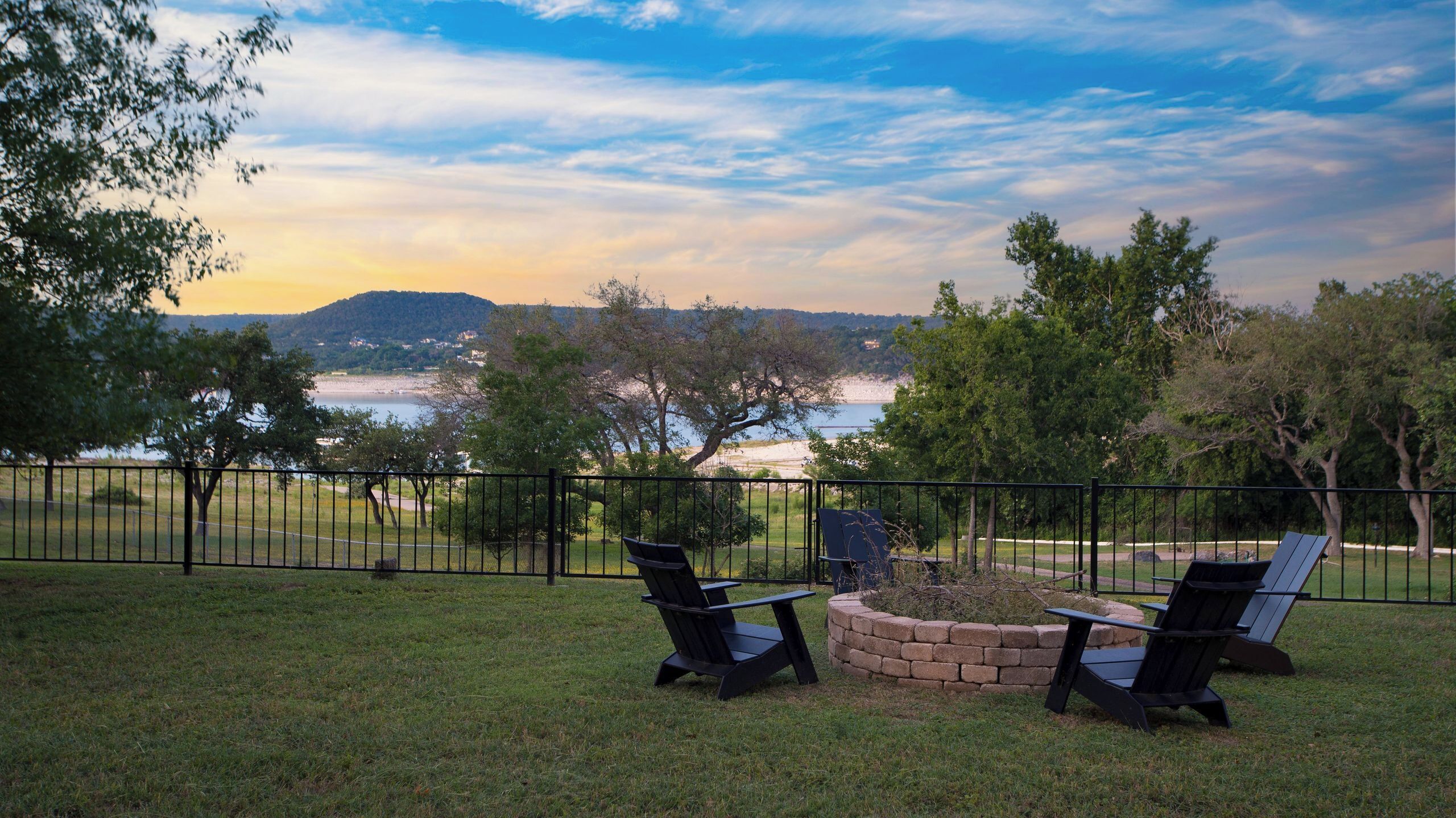 A grassy outdoor area with a stone fire pit and two Adirondack chairs, overlooking a body of water and distant hills.