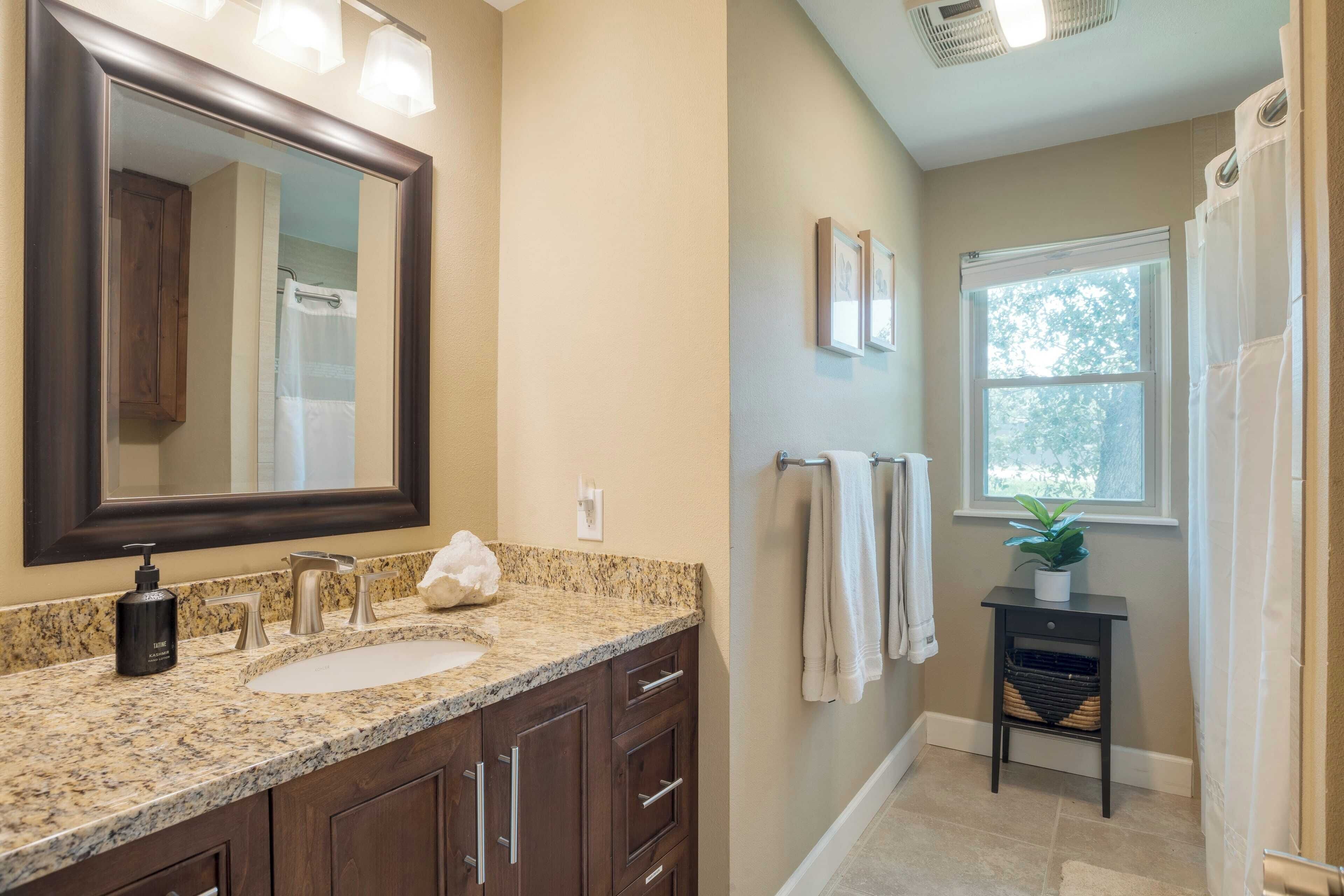 A bathroom with a granite countertop sink, a large mirror, and a window with a view of trees.