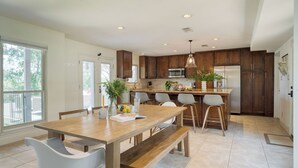 A modern kitchen with wooden cabinets, a central island, bar stools, and a dining table with chairs. - Travis by Avantstay Waterfront Lake Retreat (Austin)