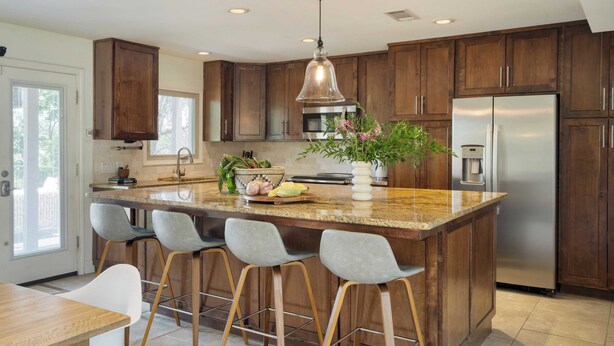 A modern kitchen with wooden cabinets, a stainless steel refrigerator, a granite island with bar stools, and a hanging pendant light. - Travis by Avantstay Waterfront Lake Retreat (Austin)