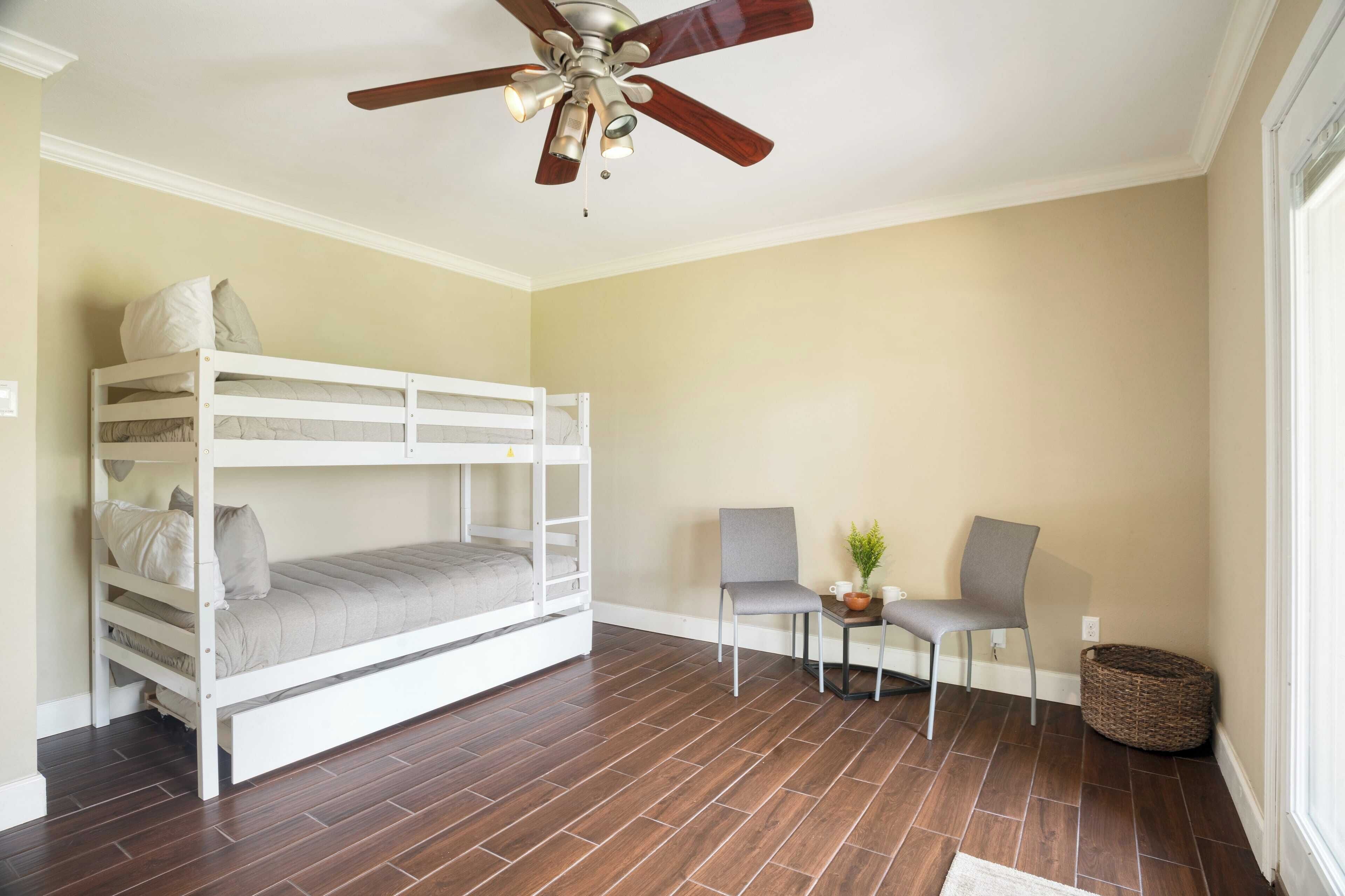 Una habitación con una cama litera, dos sillas, un ventilador de techo y una mesita con una planta.