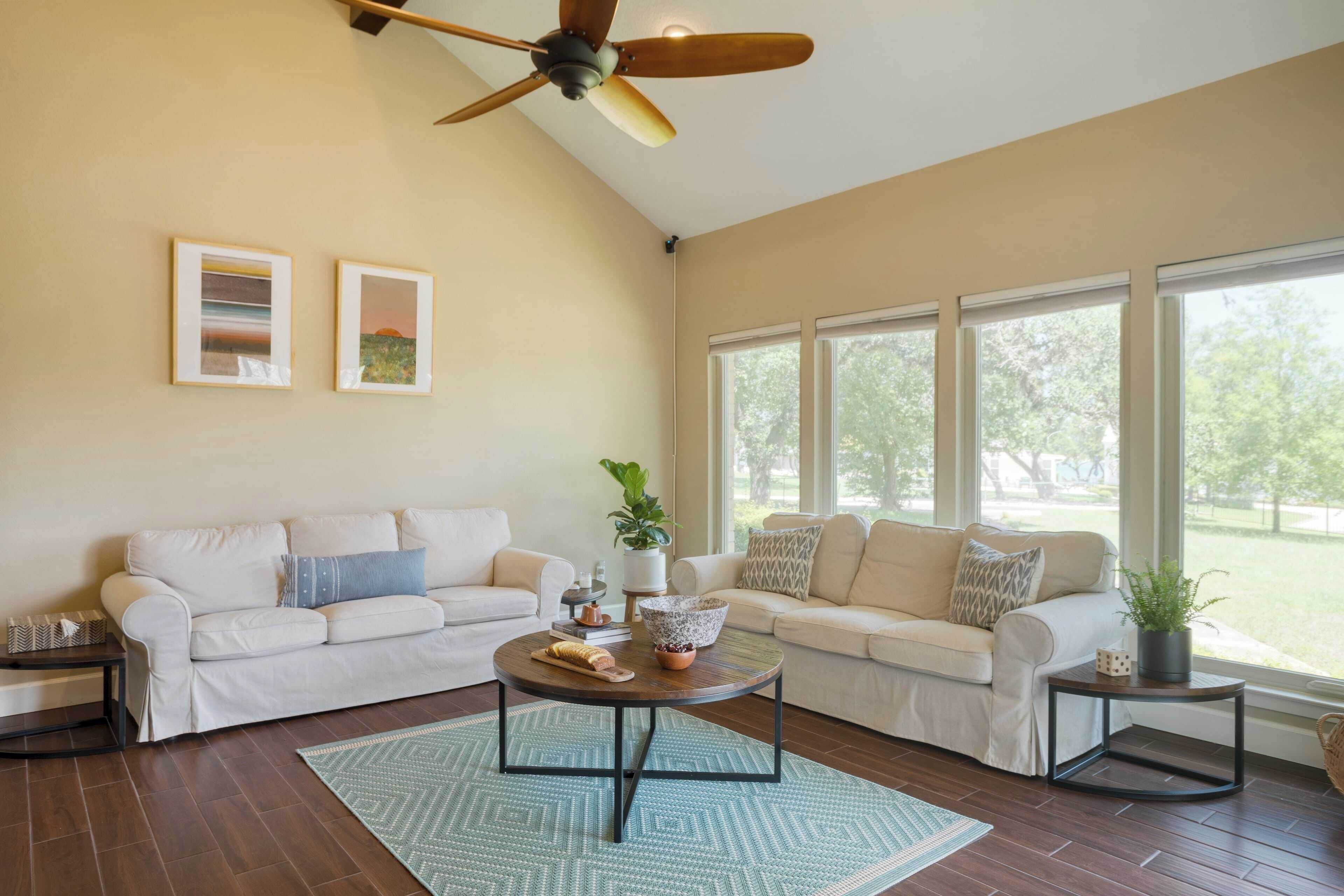 A spacious living room with two sofas, a coffee table, and a ceiling fan. Large windows provide natural light and a view of the outdoors.