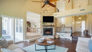 A spacious living area with a stone fireplace, a ceiling fan, and a large TV mounted on the wall. - Travis by Avantstay Waterfront Lake Retreat (Austin)