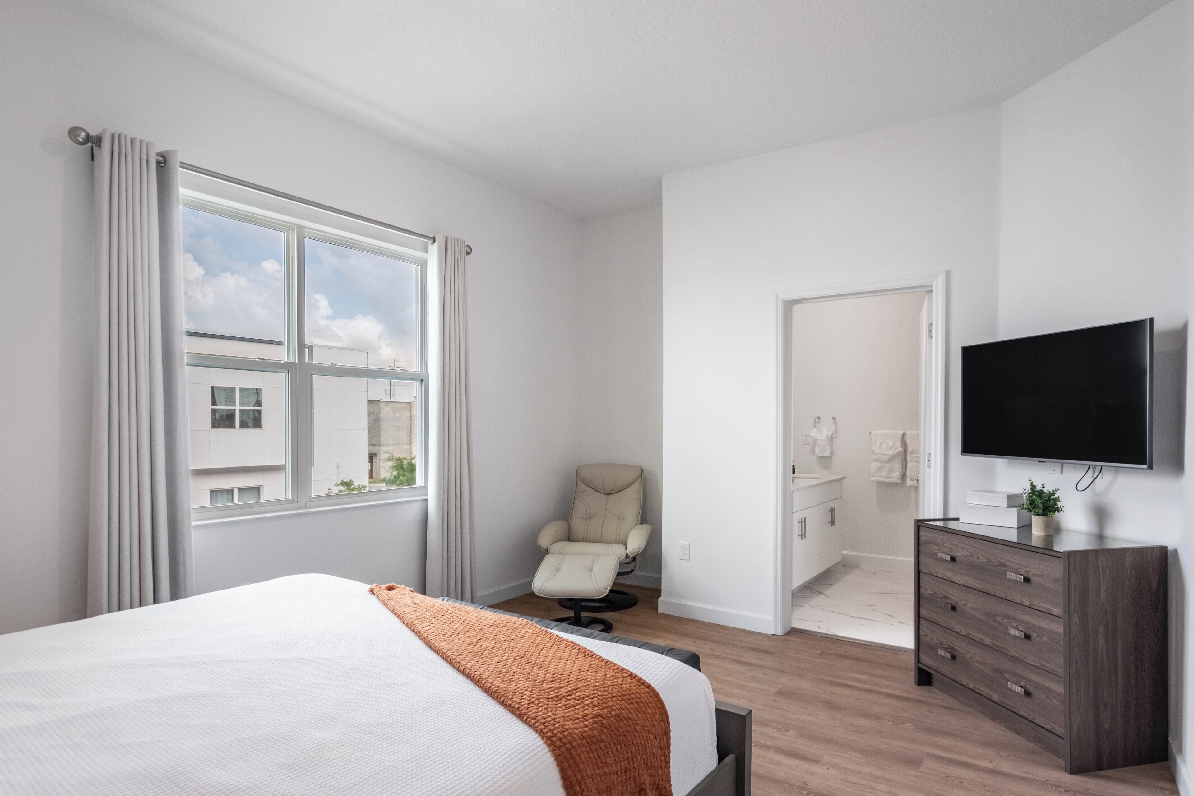 A modern hotel room with a large bed, a massage chair, a TV, and a view of the city.