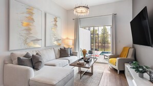 A modern living room with a sofa, coffee table, and a TV. There are abstract paintings on the wall and a chandelier.