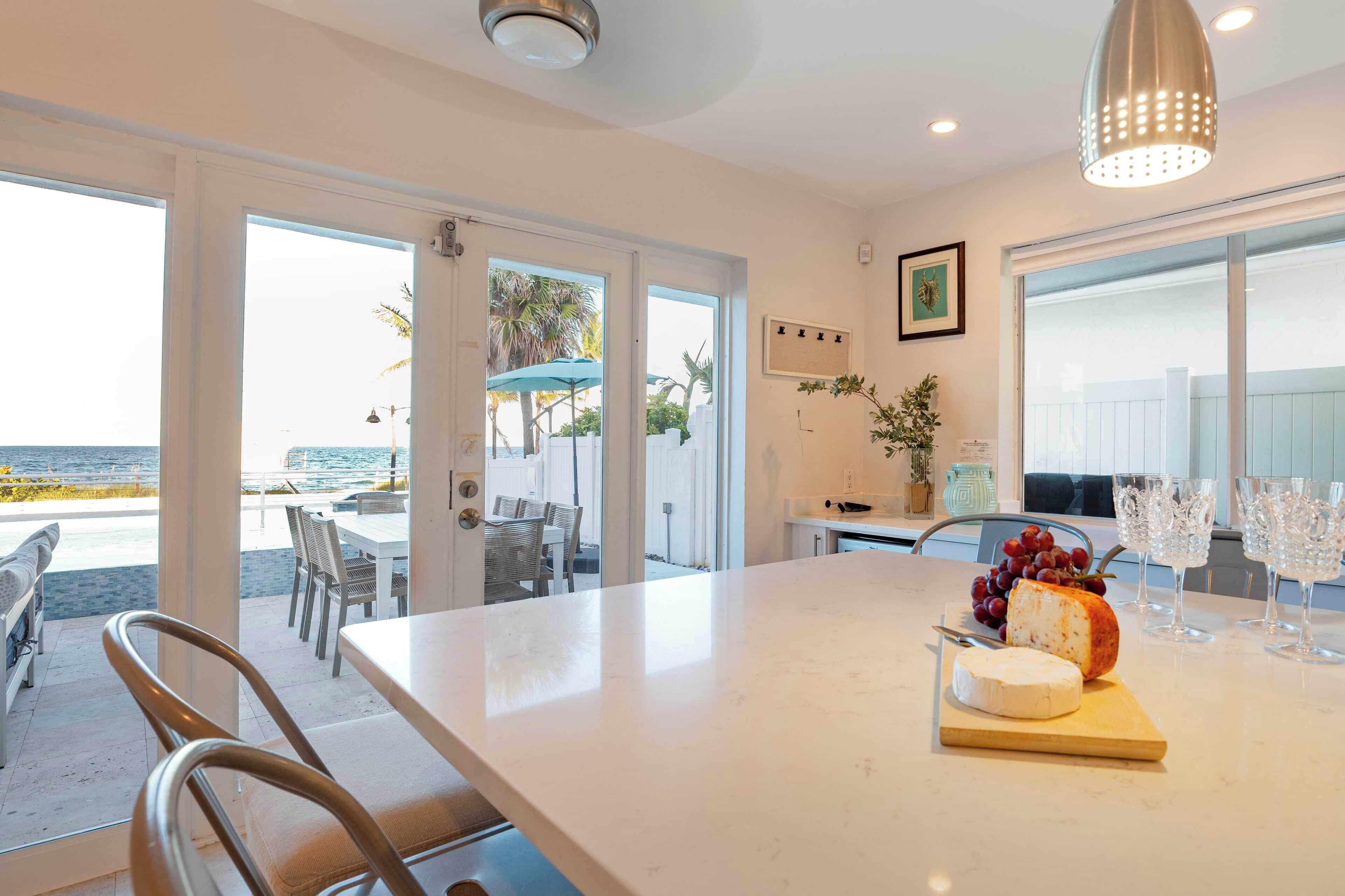 Una cocina con encimera de mármol, una presentación de quesos y uvas, y vistas a la playa a través de amplias ventanas.