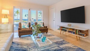 A living room with a large TV, two sofas, a coffee table with decorative items, and a view of the ocean through large windows.