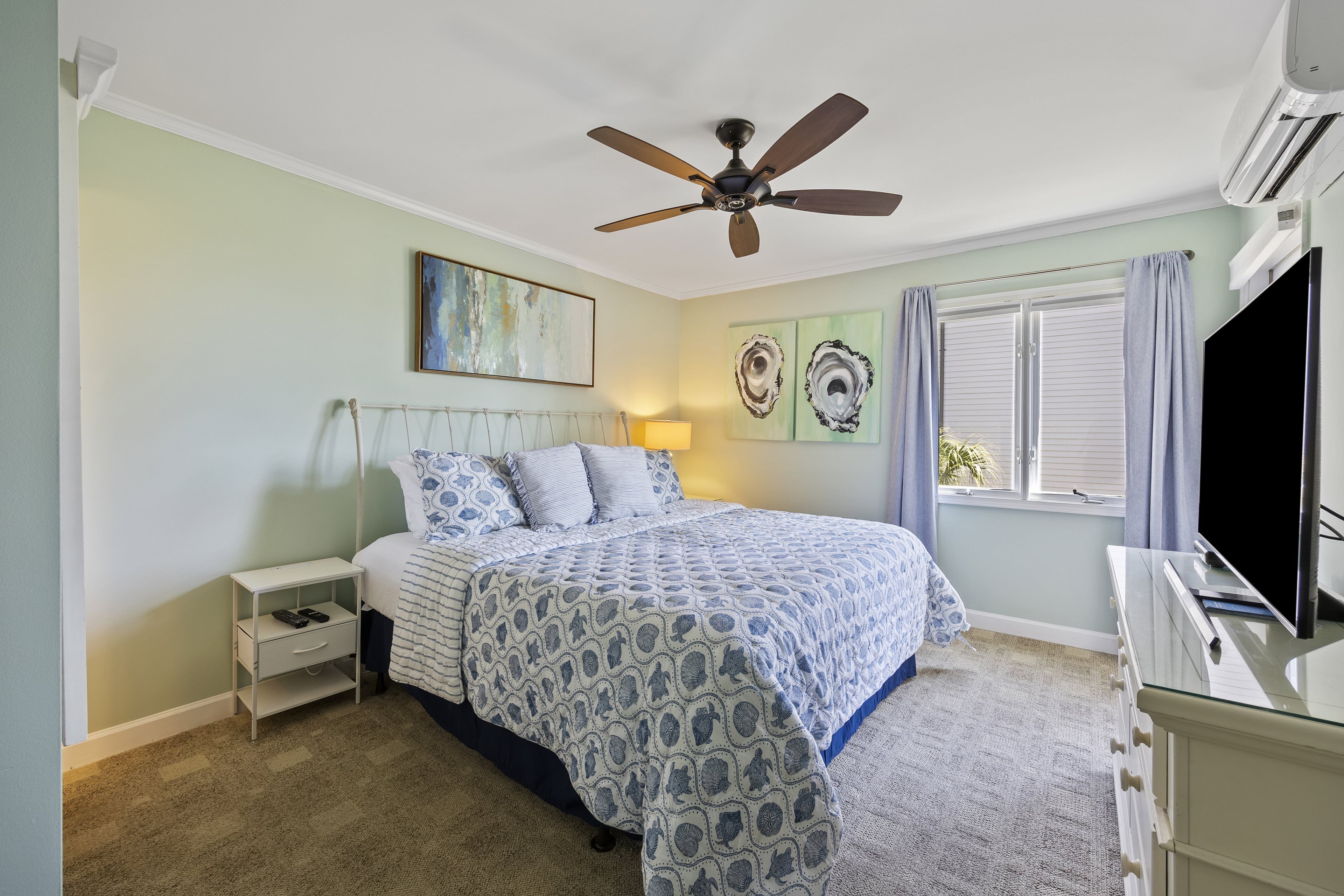 A bedroom with a bed, nightstand, TV, ceiling fan, and artwork on the walls.
