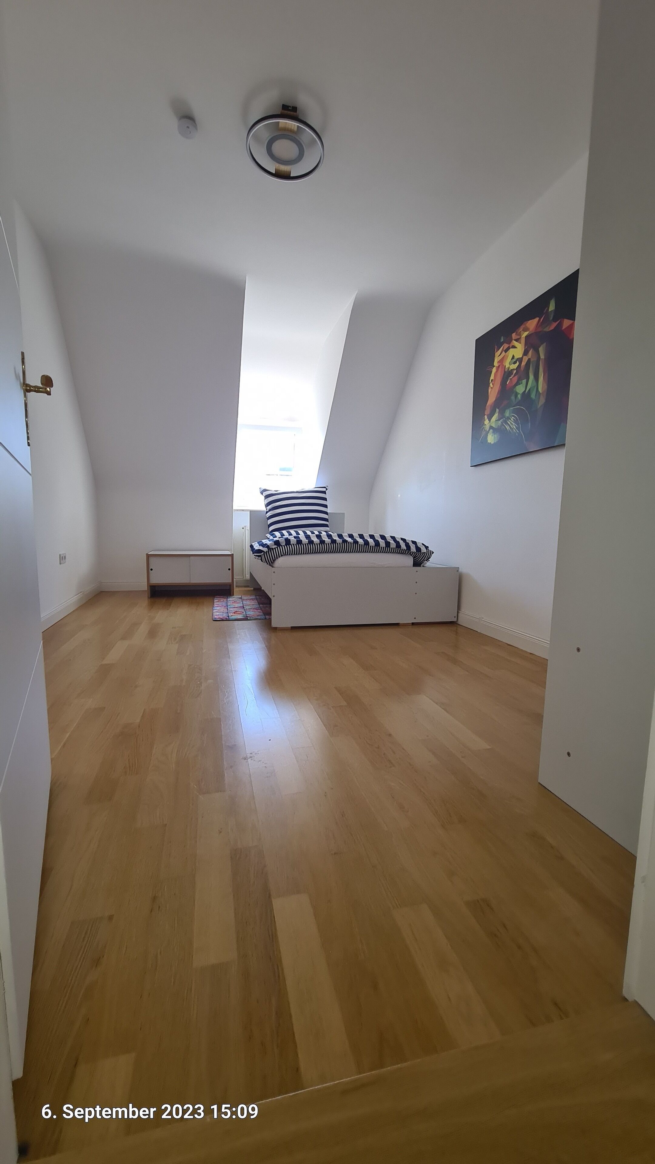 A room with a sloped ceiling, a bed with a striped pillow, a painting on the wall, and a wooden floor.