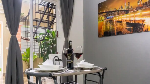 A dining area with a table set for two, a bottle of wine, and a glass of red wine. There is a painting of a cityscape on the wall.