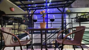 A table with two wicker chairs, a potted plant, and two wine glasses on a balcony with a metal railing and blue lighting.