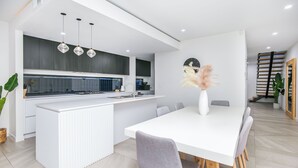 A modern kitchen with a central island, a dining area with a white table and grey chairs, and a staircase leading to an upper level.