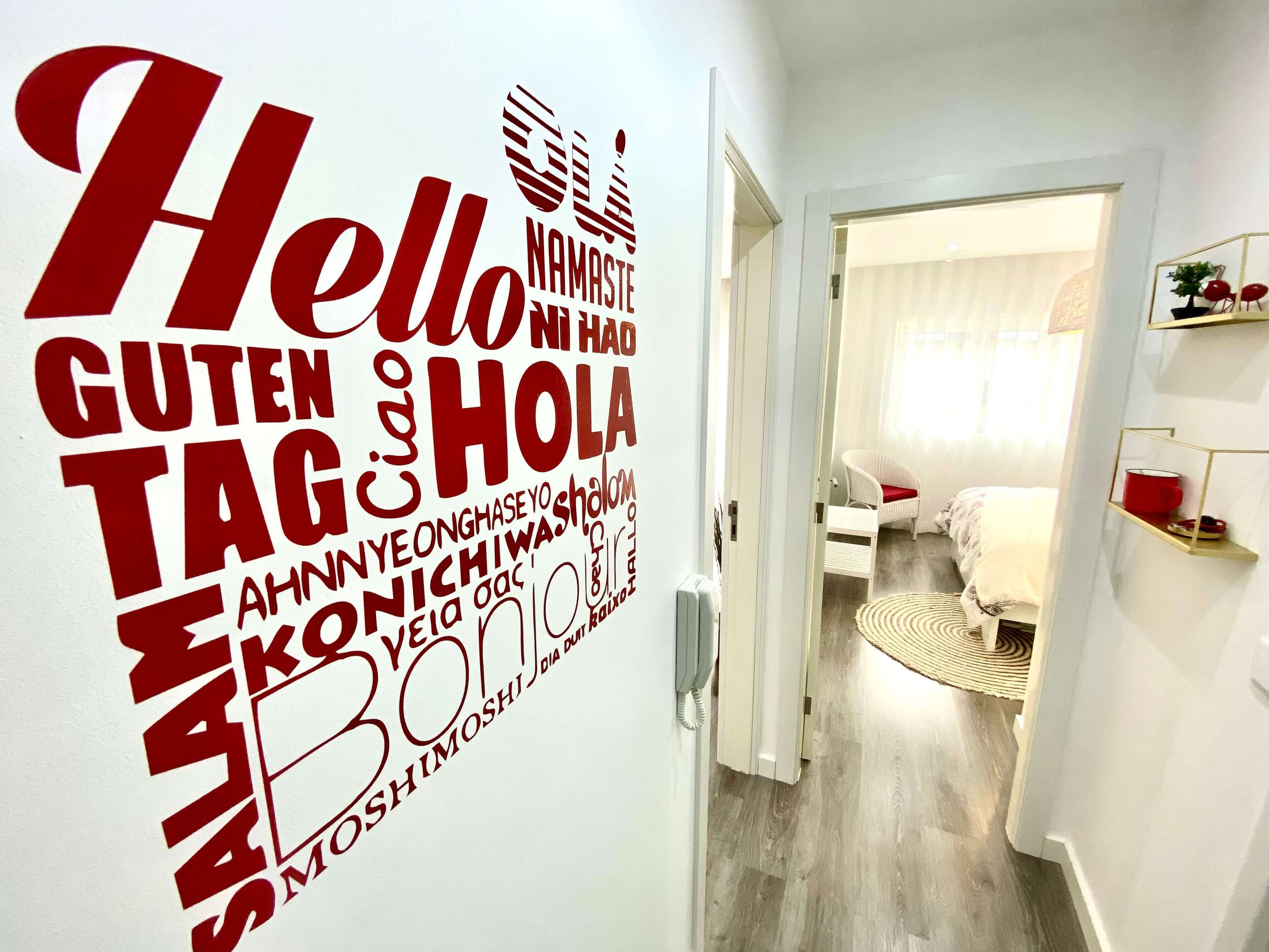 A hallway with a wall featuring multilingual 'Hello' signs and a glimpse of a bedroom with a bed, a chair, and a small table.