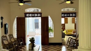 Un hall de hotel con piso a cuadros, dos puertas de madera con detalles de metal decorativo, una mesa con un jarrón y un sofá amarillo.