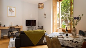 Un salon chaleureux avec un canapé, une table à manger dressée pour deux personnes, un téléviseur et une grande fenêtre donnant sur un paysage verdoyant.