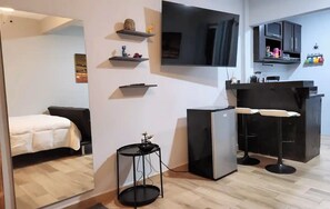 Un espace de vie compact comprenant un lit, une petite table ronde, un réfrigérateur, un téléviseur fixé au mur et un coin cuisine avec un tabouret de bar.