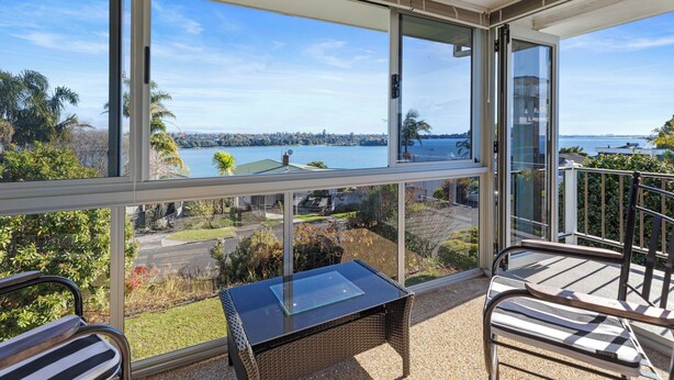 A balcony with a glass table and chairs, overlooking a body of water and a cityscape.