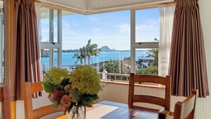 A dining area with a table and chairs, a vase with flowers, and a view of the sea and a mountain through large windows.